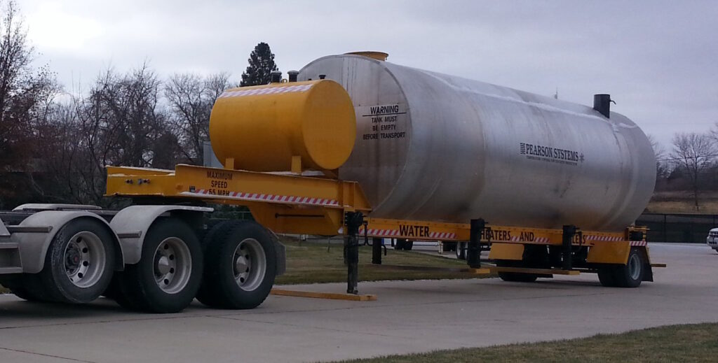 A large cylindrical Pearson portable water storage tank and water heating system labeled WATER is mounted on a yellow trailer attached to a truck. The tank has safety warnings and is situated outdoors on a paved driveway with grass and trees nearby.