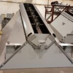 Large, industrial aggregate bin with support structures sits inside a spacious RexCon factory with a high ceiling and yellow overhead crane in the background. The aggregate batcher or scale has an angled, open-top design.