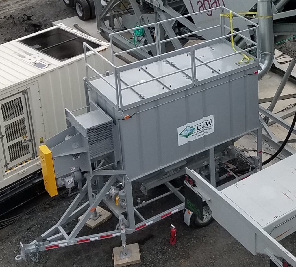 A large, industrial gray machine with a C&W company logo, metal railing on top, and an angled chute stands on a construction site surrounded by other equipment.