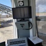 A CommandBatch Command Alkon Control panel setup inside a small booth with a monitor, gauge, and RexCon equipment. Outside the window, industrial conveyor belts and structures are visible under a sunny sky.
