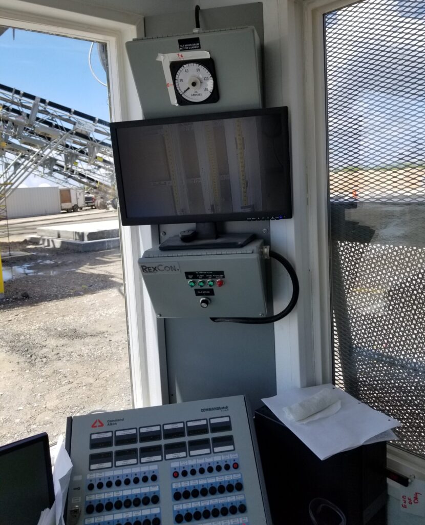 A CommandBatch Command Alkon Control panel setup inside a small booth with a monitor, gauge, and RexCon equipment. Outside the window, industrial conveyor belts and structures are visible under a sunny sky.