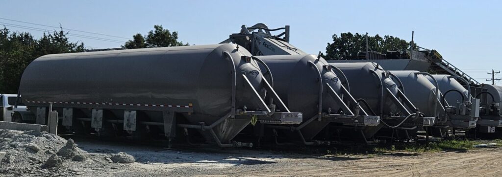 Several large silver cement trailers known as cement pigs, cement guppies, or horizontal cement storage tanks are parked in a row on a dirt lot, with construction equipment and trees in the background under a clear blue sky.