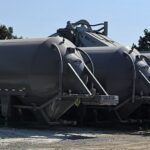 Several large silver cement trailers known as cement pigs, cement guppies, or horizontal cement storage tanks are parked in a row on a dirt lot, with construction equipment and trees in the background under a clear blue sky.