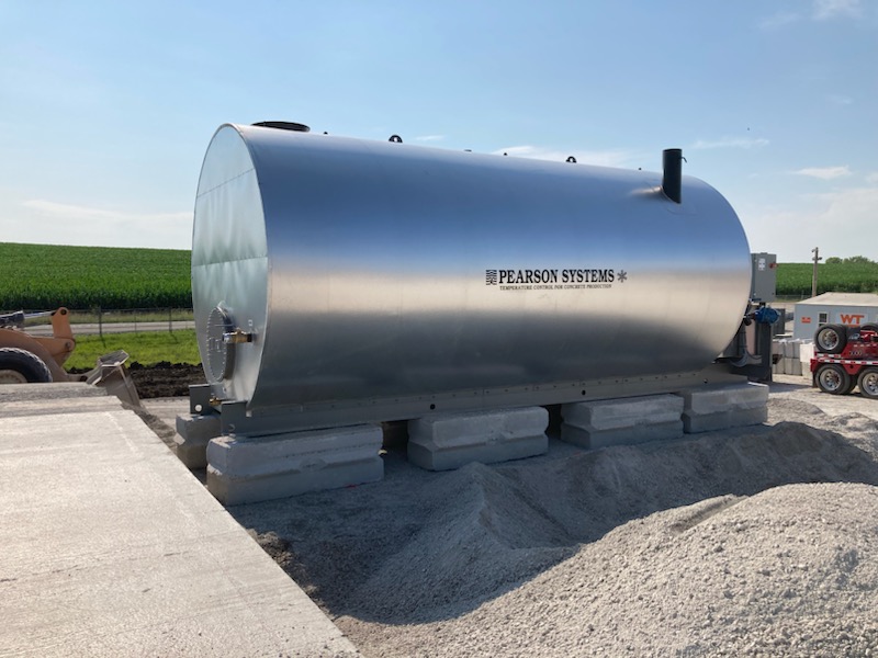 A large, cylindrical metal tank labeled Pearson Systems sits on concrete supports outdoors, surrounded by gravel and construction materials, with green fields and blue sky in the background.