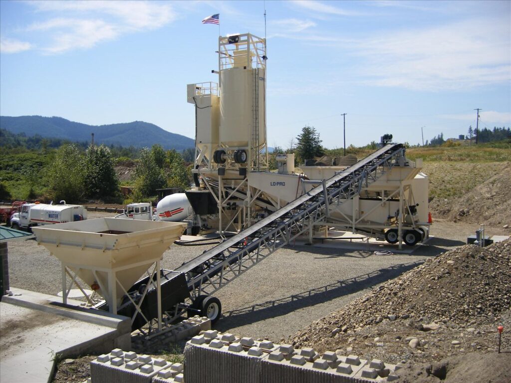 A concrete batching plant with silos, conveyors, and hoppers sits on a gravel lot under a partly cloudy sky, with vehicles and hills visible in the background. An American flag flies atop the central structure.