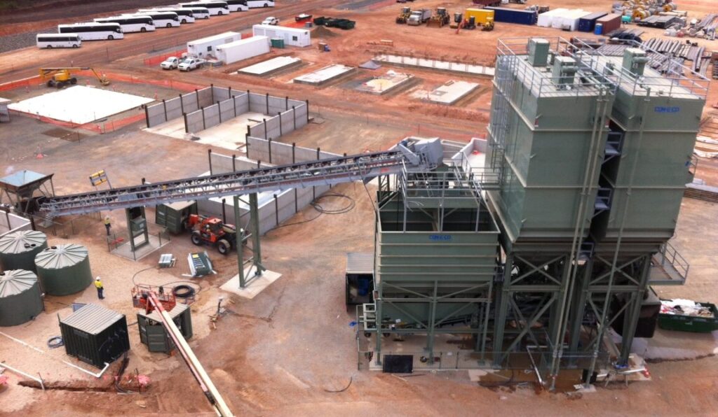 Aerial view of an industrial concrete plant site with a conveyor feeding a turnhead, construction vehicles, and workers surrounded by dirt and building materials.