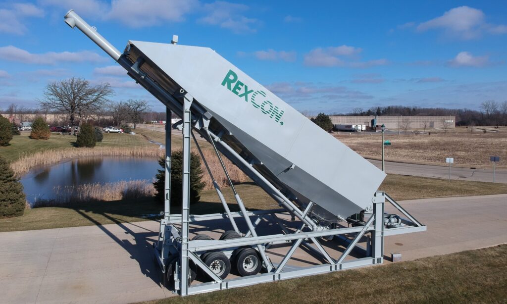 Large, silver industrial machine labeled RexCon mounted on a trailer, tilted upward on a paved road with grassy fields, trees, and a pond in the background under a blue sky.