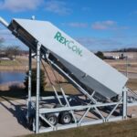 Large, silver industrial machine labeled RexCon mounted on a trailer, tilted upward on a paved road with grassy fields, trees, and a pond in the background under a blue sky.