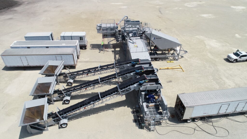 Aerial view of an industrial mobile central mix CONECO concrete plant with multiple conveyor belts, surrounded by trailers, containers, and a white pickup truck on a dirt lot.
