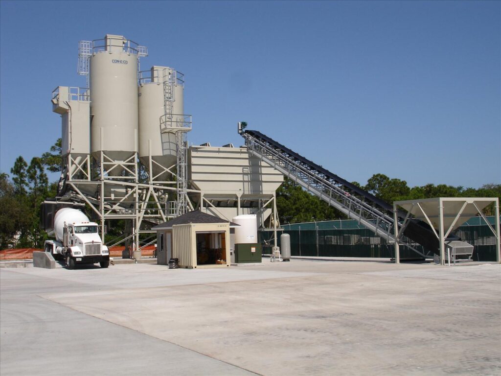 A CON-E-CO concrete batching plant with large silos, conveyor belts, a concrete mixer truck, and small utility buildings on a clean, paved lot, surrounded by fencing and trees under a clear blue sky.