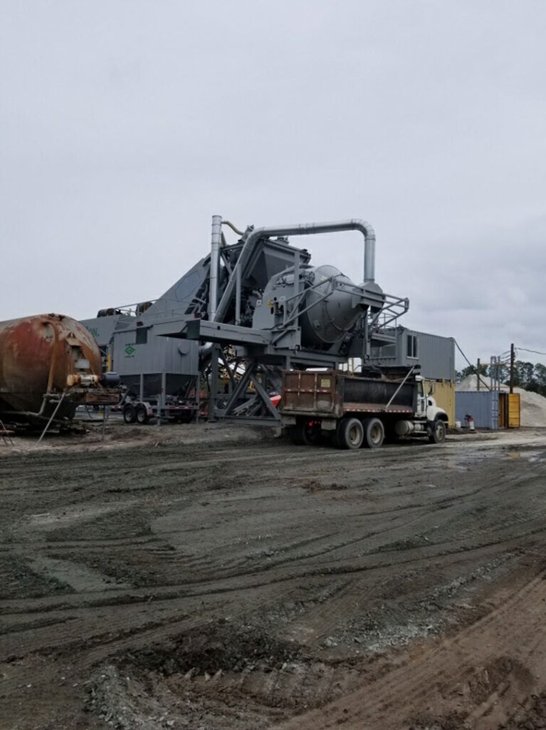 A large industrial central mix concrete paving plant wth a RexCon tilt mixer stands on a gravel lot under an overcast sky, with a dump truck parked in front and piles of materials nearby.