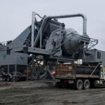 A large industrial central mix concrete paving plant wth a RexCon tilt mixer stands on a gravel lot under an overcast sky, with a dump truck parked in front and piles of materials nearby.