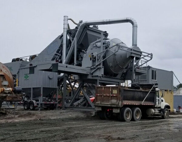 A large industrial central mix concrete paving plant wth a RexCon tilt mixer stands on a gravel lot under an overcast sky, with a dump truck parked in front and piles of materials nearby.