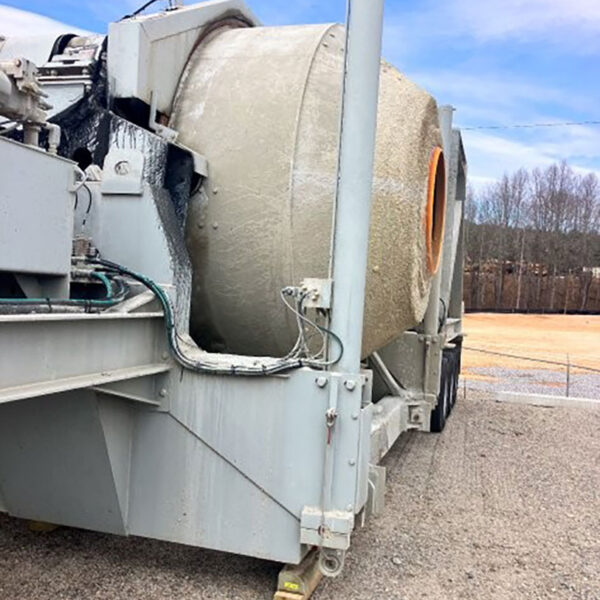 A close-up side view of a large concrete mixer truck, showing the rotating drum coated with dried cement, parked on a gravel lot with trees and a cloudy sky in the background.