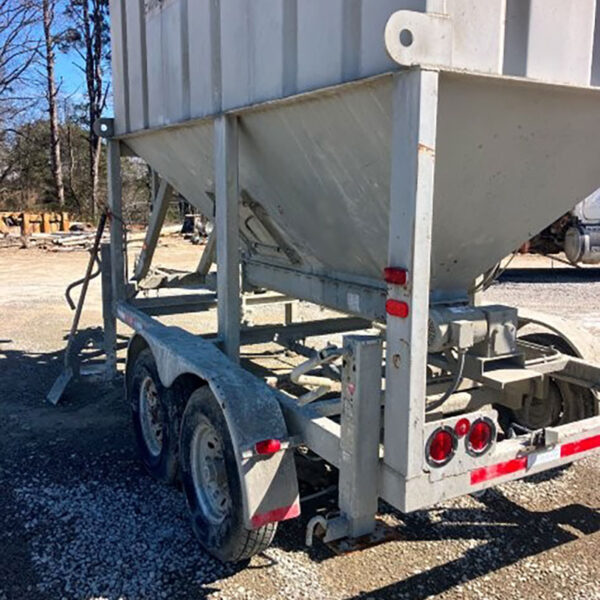 A close-up view of a metal trailer with a large hopper mounted on it, parked on gravel. The trailer has dual wheels and red brake lights. Trees and construction materials are visible in the background.