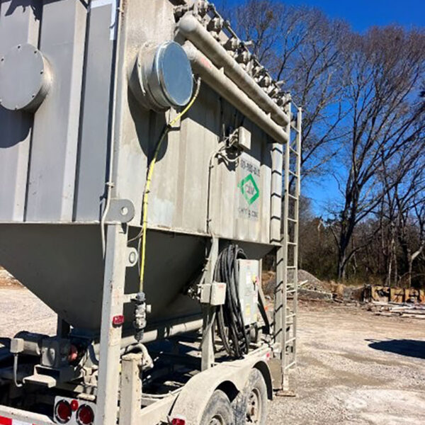 Large industrial equipment mounted on a double-axle trailer sits outdoors on a gravel lot. The unit is gray, has pipes, hoses, and a ladder on the side, with trees and a clear blue sky in the background.