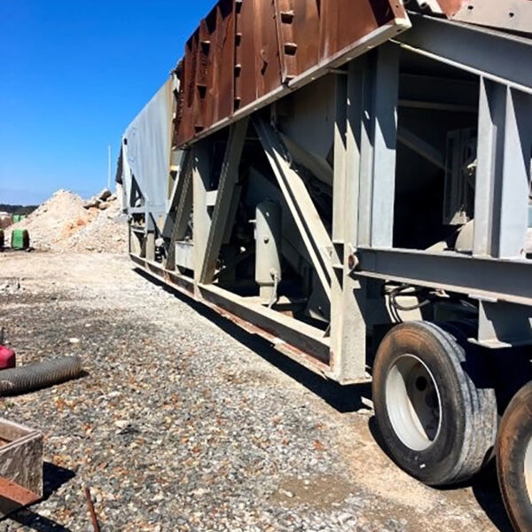 Large industrial machine with metal frame and rusted sections sits on wheels at a rocky construction site under a clear blue sky. Piles of gravel and construction materials are visible in the background.