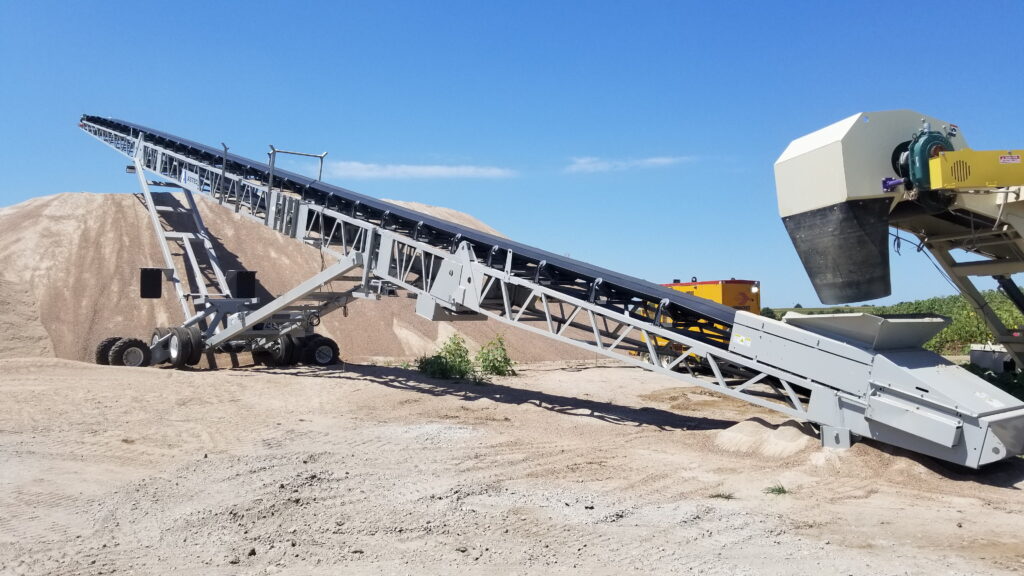 A long industrial conveyor belt system transports sand to create a large pile at an outdoor site, with a blue sky and some machinery in the background.