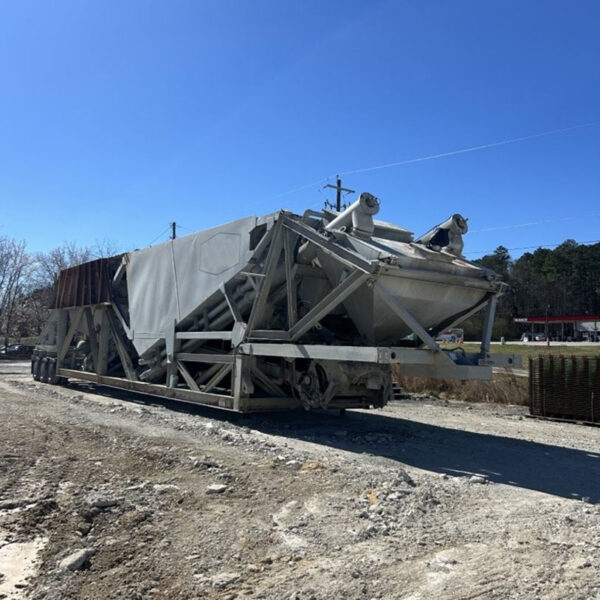 A large, industrial metal structure or machine sits on a dirt road under a clear blue sky, with trees and utility poles in the background.