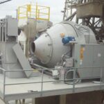 A large industrial concrete plant with a CONECO Tilt mixer or sits on a metal platform with railings. Nearby are control panels, a small ladder, and equipment. The background shows an outdoor, industrial setting.