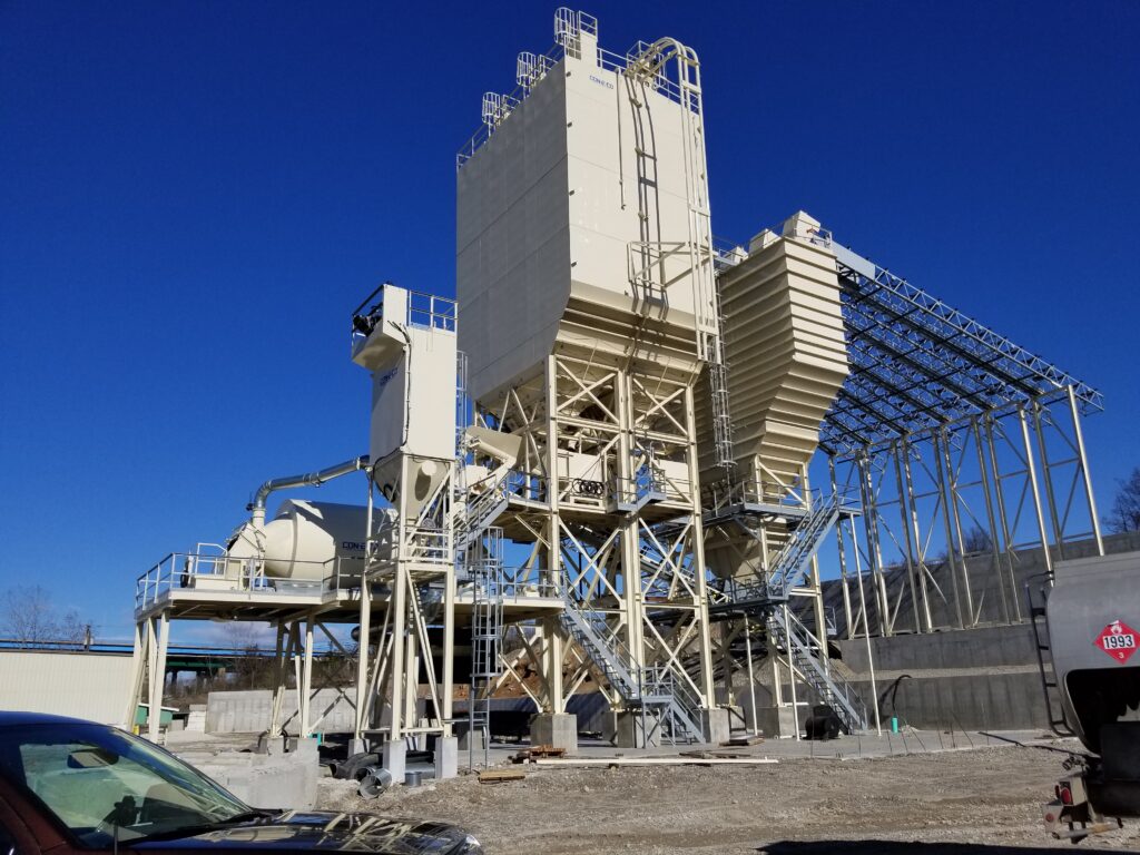 A large industrial concrete batching plant with multiple tall silos, support structures, and staircases stands under a clear blue sky in an outdoor construction area. A vehicle is partially visible in the foreground.