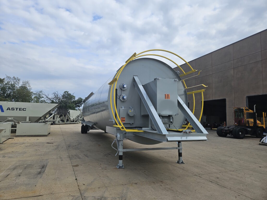 A large cylindrical metal industrial tank known as a Vertical Pig or Vertical Guppy or Vertical Keep Full SIlo with yellow safety railings stands on supports outdoors near a warehouse, with construction equipment and other machinery visible in the background.