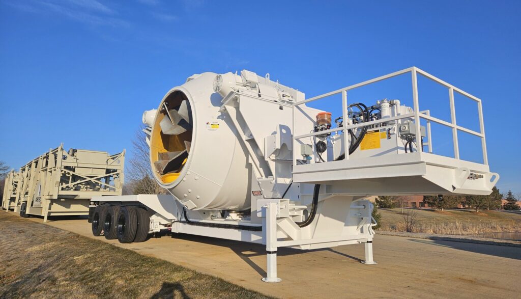 A large white industrial mobile concrete mixer is mounted on a trailer with multiple wheels and positioned on a paved area under a clear blue sky.