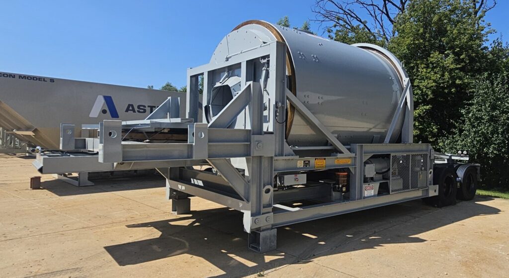 A large RexCon Concrete Horizontal Shrink Mixer is mounted on a trailer platform outdoors, with trees and a clear blue sky in the background. An Aggregate Bin is visible nearby.