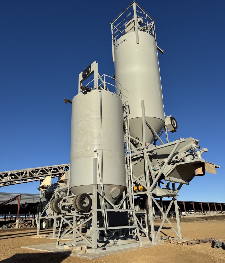 Two large, light gray industrial portable cement silos stand atop and nearby a concrete plant with ladders and railings are set on a metal framework at a construction site, with conveyor belts and surrounding steel structures under a clear blue sky. A shadow of the photographer is visible.