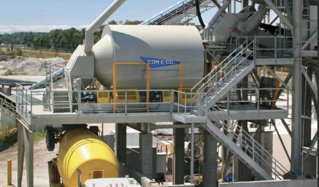 An industrial concrete mixer HRM Horizontal Reversing MIXER labeled CON-E-CO is positioned on a metal platform with stairs and railings, surrounded by machinery and a yellow tank at a concrete production facility.