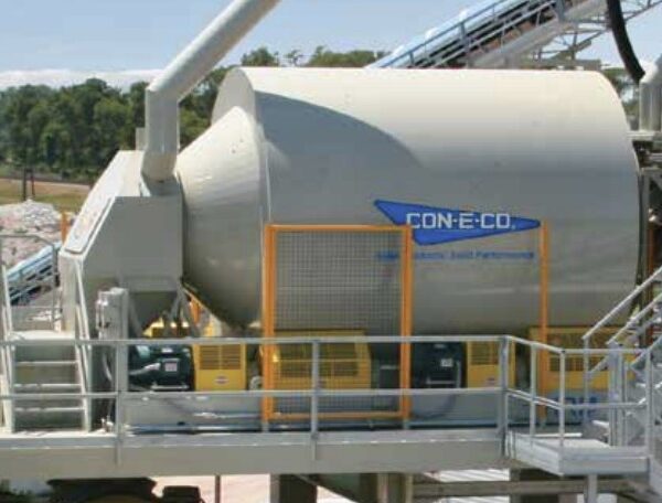 An industrial concrete mixer HRM Horizontal Reversing MIXER labeled CON-E-CO is positioned on a metal platform with stairs and railings, surrounded by machinery and a yellow tank at a concrete production facility.