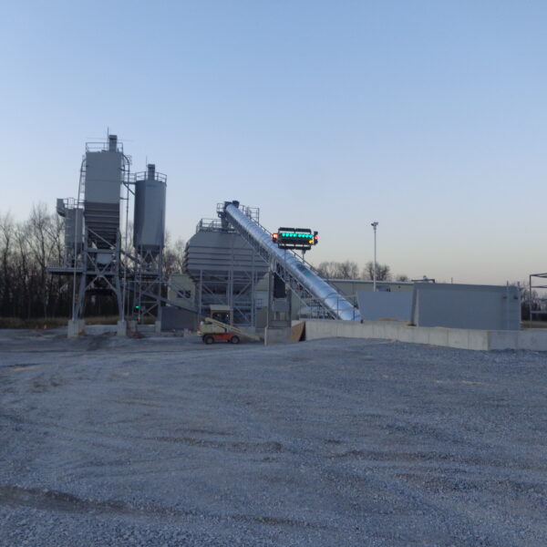 Industrial concrete batching plant with silos, conveyor, and structures on a gravel lot; bare trees and clear sky in the background.