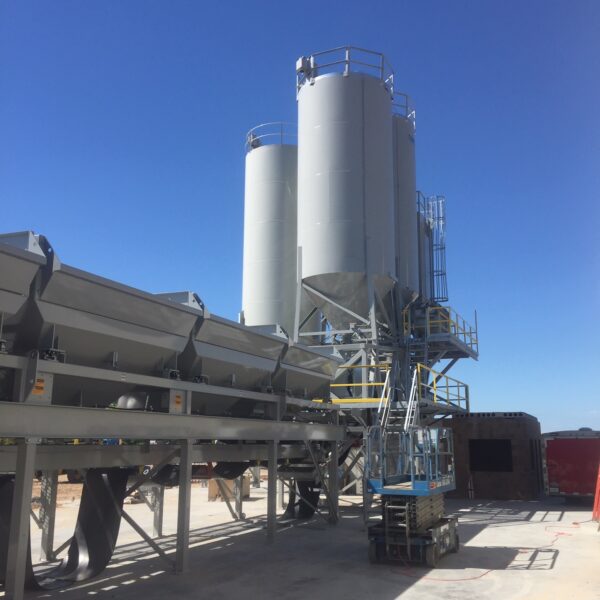 Large industrial silos and metal equipment under a clear blue sky, with platforms, ladders, and walkways, set on a concrete surface at an outdoor industrial site.