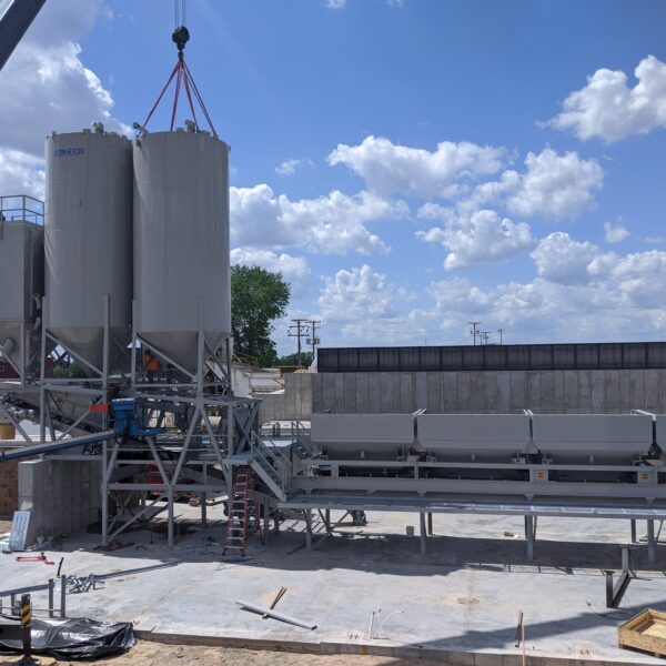 A construction site features large vertical silos being installed by a crane, with a conveyor system and workers present. The sky is blue with scattered clouds, and various equipment and materials are visible on the ground.