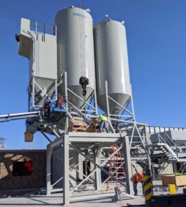Two workers in safety gear are assembling or maintaining industrial concrete batch plant equipment near large vertical storage cement silos at a construction site, using ladders and scaffolding on a sunny day.