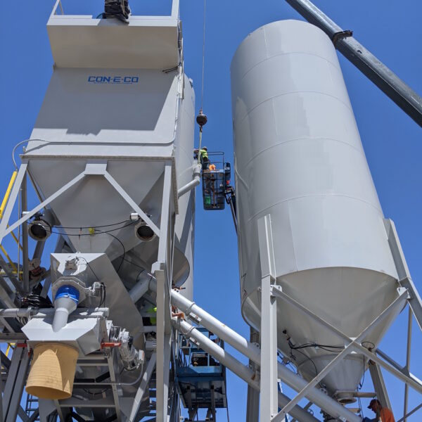 Two workers in safety gear work on large industrial silos and a concrete plant machinery under a clear blue sky, using a crane and scaffolding for access and support.