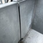 A black cable extends vertically from a small metal junction box mounted on the side of a large, empty, galvanized metal container with visible seams and a small hole at the bottom.