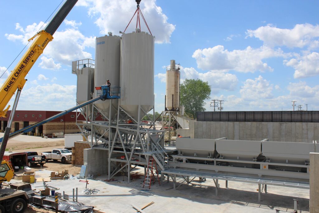 A crane lifts a large cylindrical tank onto a metal structure at an industrial construction site of a concrete batch plant, while a worker on a lift assists. Buildings, vehicles, and equipment are visible under a partly cloudy sky.