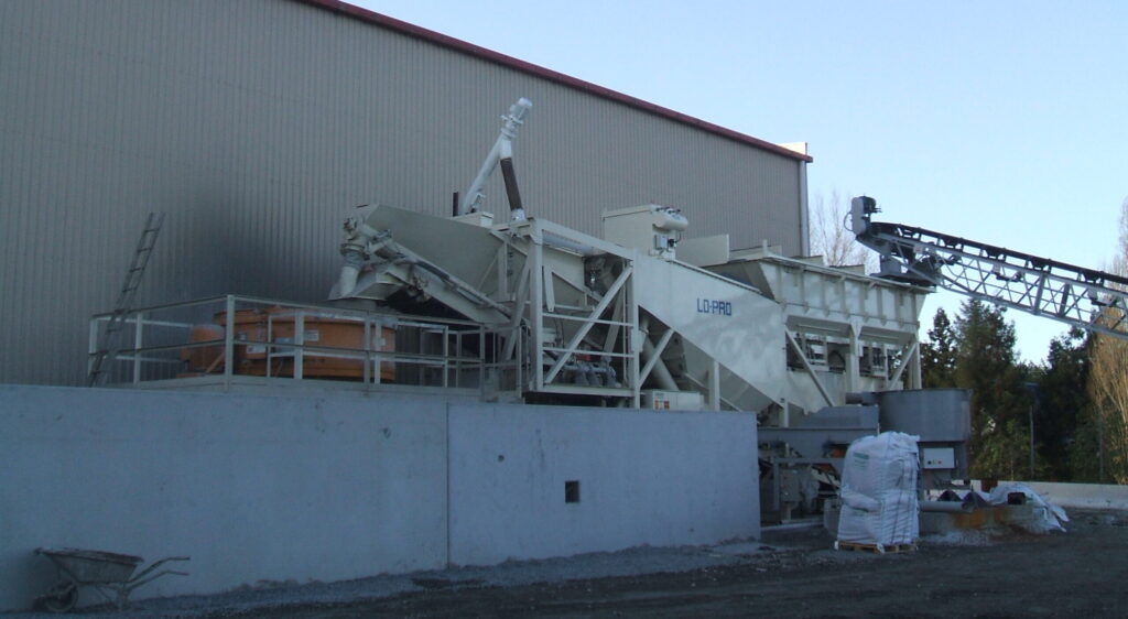 A large industrial concrete batching plant with pan mixer hoppers, conveyor belts, and mixing machinery stands next to a tall building. Pallets of materials and bags are stacked nearby under a clear sky.