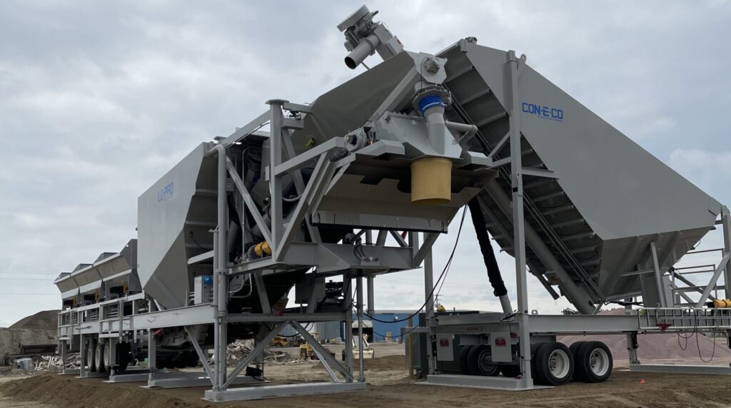 Large industrial concrete batching plant with 450 bbl metal framework and conveyor belts set up outdoors on a dirt lot under a cloudy sky. The equipment is labeled CON-E-CO and a truck is parked beside it.
