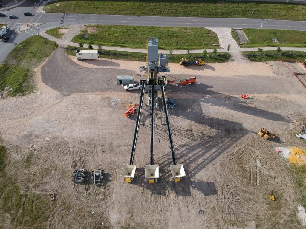 Aerial view of an industrial construction site consisting of a Central Mix Wet Batch RexCon Model S concrete batch plant or concrete paving plant with three conveyor belts extending from machinery, vehicles, and equipment scattered around, and a road running parallel at the top of the image.