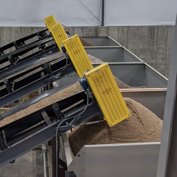 Several conveyor belts with yellow safety guards transport granular material into large metal storage bins inside an industrial facility with concrete walls and a high, white ceiling.