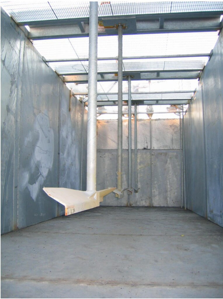 A large, empty industrial metal tank with a grated roof, pipes, and a slanted metal chute hangs from above. The interior walls and floor show signs of wear and discoloration.