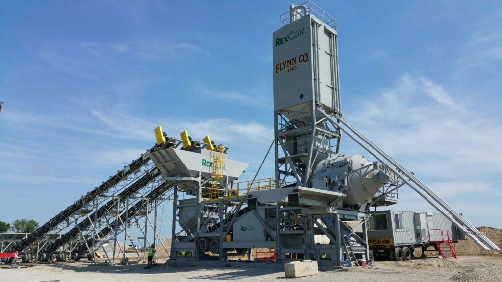 A large industrial RexCon Model S concrete batching plant with conveyor belts, silos, mixers, and machinery under a clear blue sky at a construction site. The equipment is marked with REXCON and FLYNN CO.