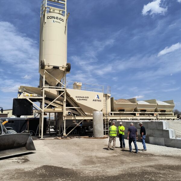 Four people in safety gear stand near a large concrete batching plant with silos and storage bins under a partly cloudy sky. Construction materials and equipment are visible around the site.