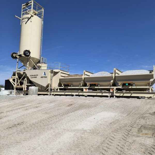 A large beige industrial concrete batching plant with multiple gravel bins and a tall silo stands on a dirt lot under a clear blue sky.