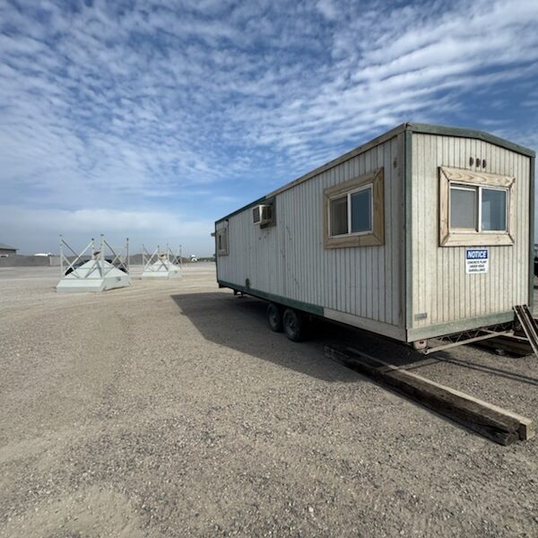 A weathered, light gray mobile concrete plant office trailer sits on gravel under a partly cloudy sky. Wooden planks lead to its door and some metal structures are visible in the background.