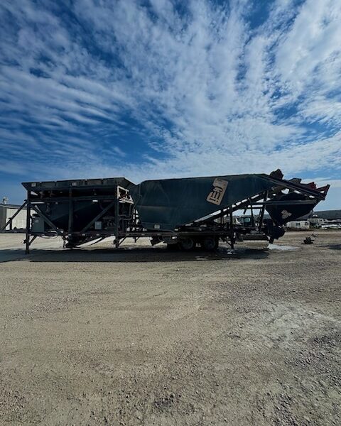 A large industrial machine, known as a portable concrete batch plant sits on a gravel lot under a blue sky with scattered clouds. The equipment is covered with a tarpaulin and flanked by industrial buildings.