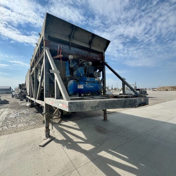A large industrial concrete plant trailer with metal support legs stands on a concrete surface under a partly cloudy sky. Machinery, including a blue air compressor, is mounted at the front of the trailer.