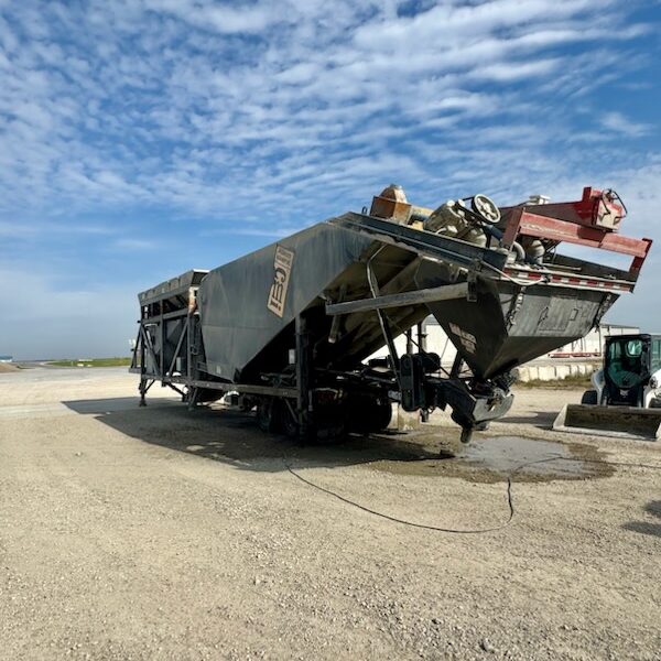 A large industrial machine, known as a concrete plant, sits on a gravel lot under a blue sky with scattered clouds. Various tools and equipment are visible on and around the machine.